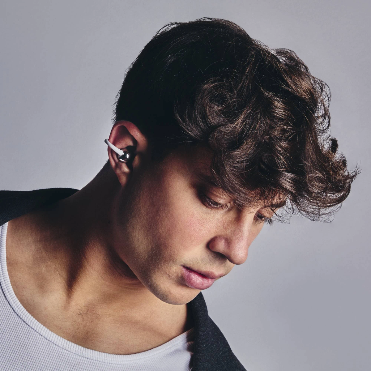 Un hombre con cabello rizado oscuro usando un auricular. Viste camiseta blanca y chaqueta negra. Está mirando hacia abajo.