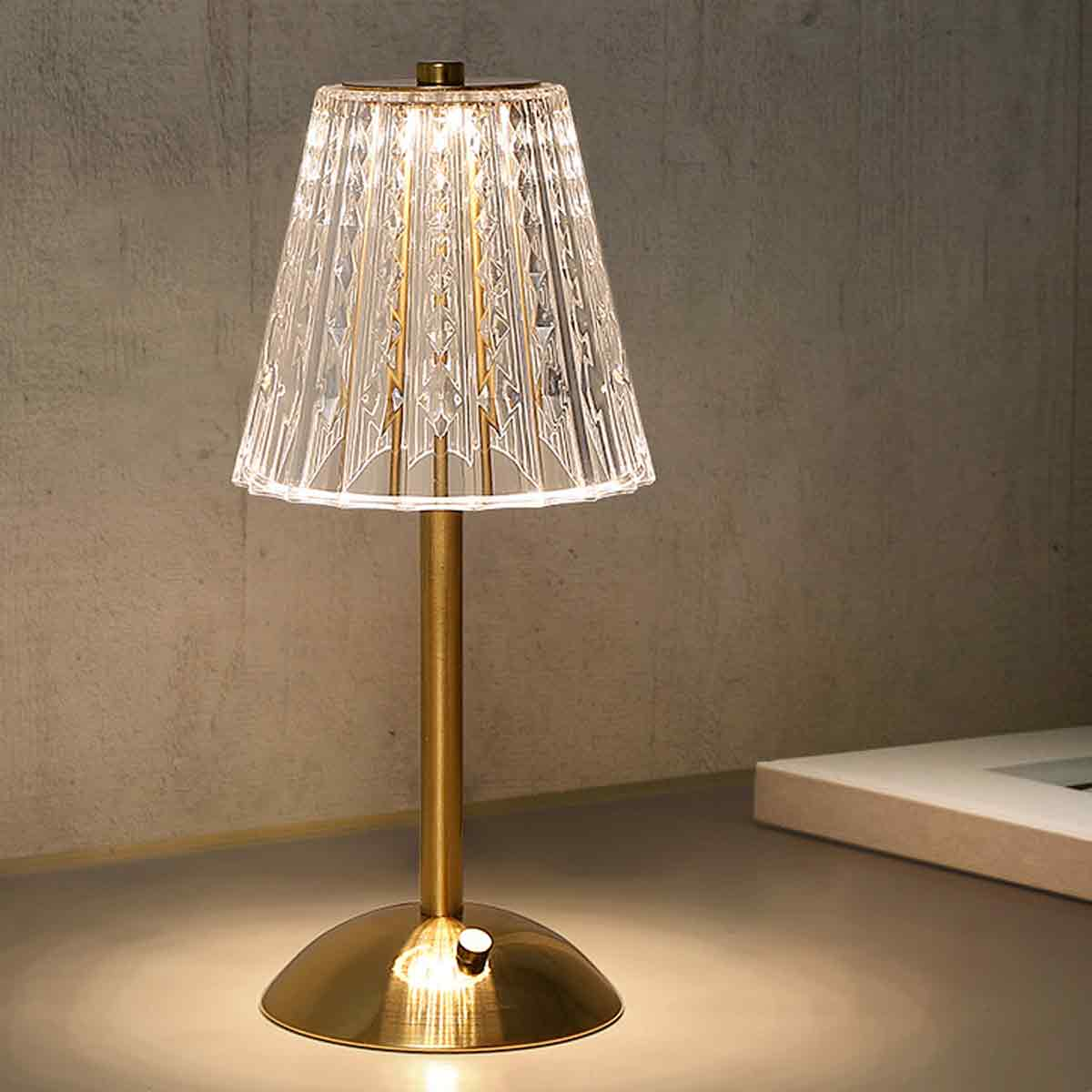 Eine goldene Tischlampe mit Kristallschirm steht auf einem Tisch. Der Hintergrund ist eine graue Wand und ein weißer Rahmen.