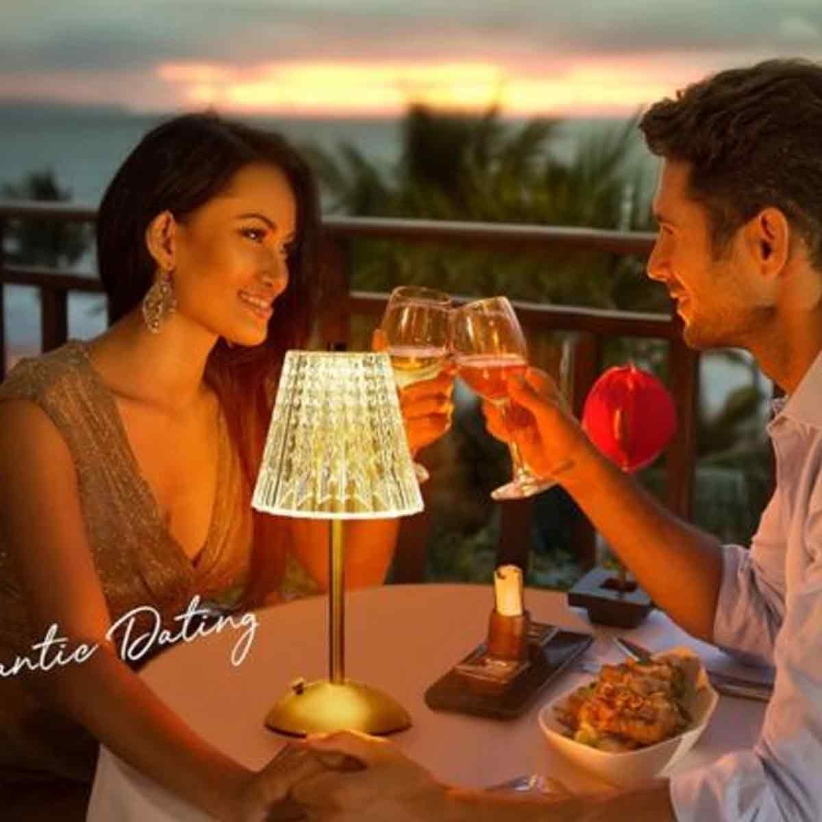 Ein Paar prostet sich mit Weingläsern an einem Tisch zu, beleuchtet von einer Lampe. Essen und eine rote Dekoration sind vorhanden.