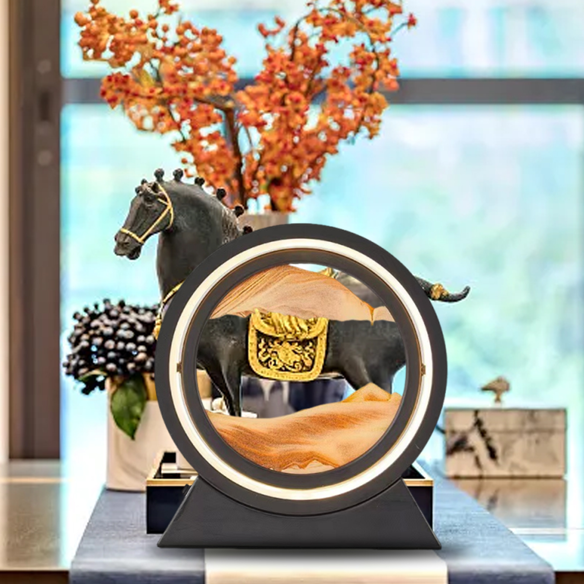 Eine dekorative Szene mit einer schwarzen Pferdestatue, einem Sandkunstwerk und orangefarbenen Blumen in einer Vase. Der Hintergrund ist unscharf.
