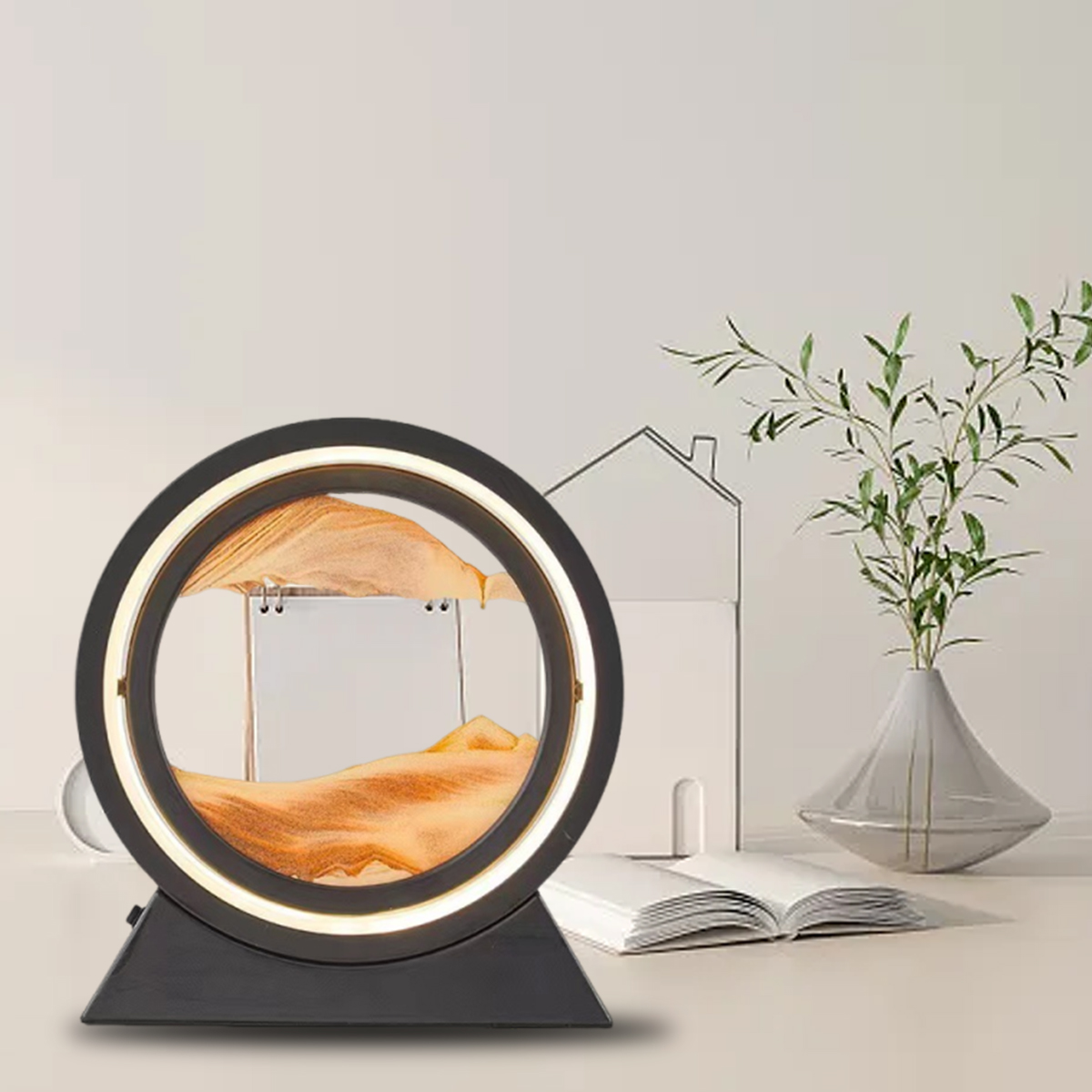 Eine Sandkunstlampe mit orangefarbenem Sand in einem runden Rahmen. Rechts befindet sich eine Vase mit grünen Pflanzen und ein offenes Buch.