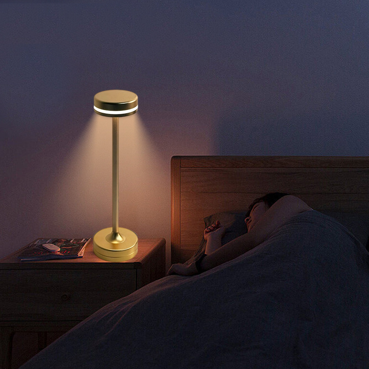 Eine goldene Nachttischlampe beleuchtet ein Bett. Eine Person schläft unter einer Bettdecke. Ein Nachttisch mit einer Zeitschrift steht am Bett.