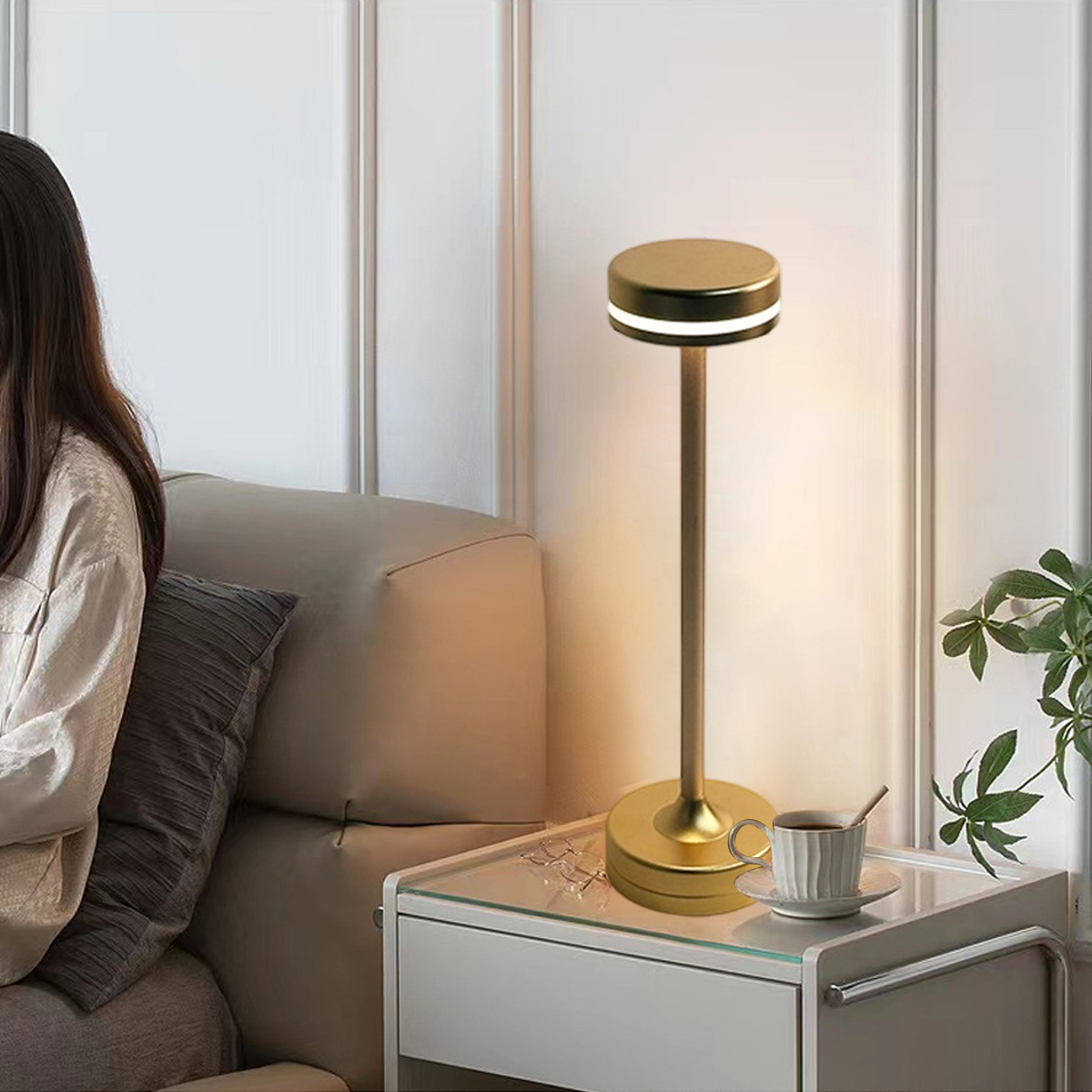Eine goldene Lampe auf einem Glastisch neben einem Sofa. Eine Tasse Kaffee und eine Pflanze stehen ebenfalls auf dem Tisch. Eine Person sitzt auf dem Sofa.