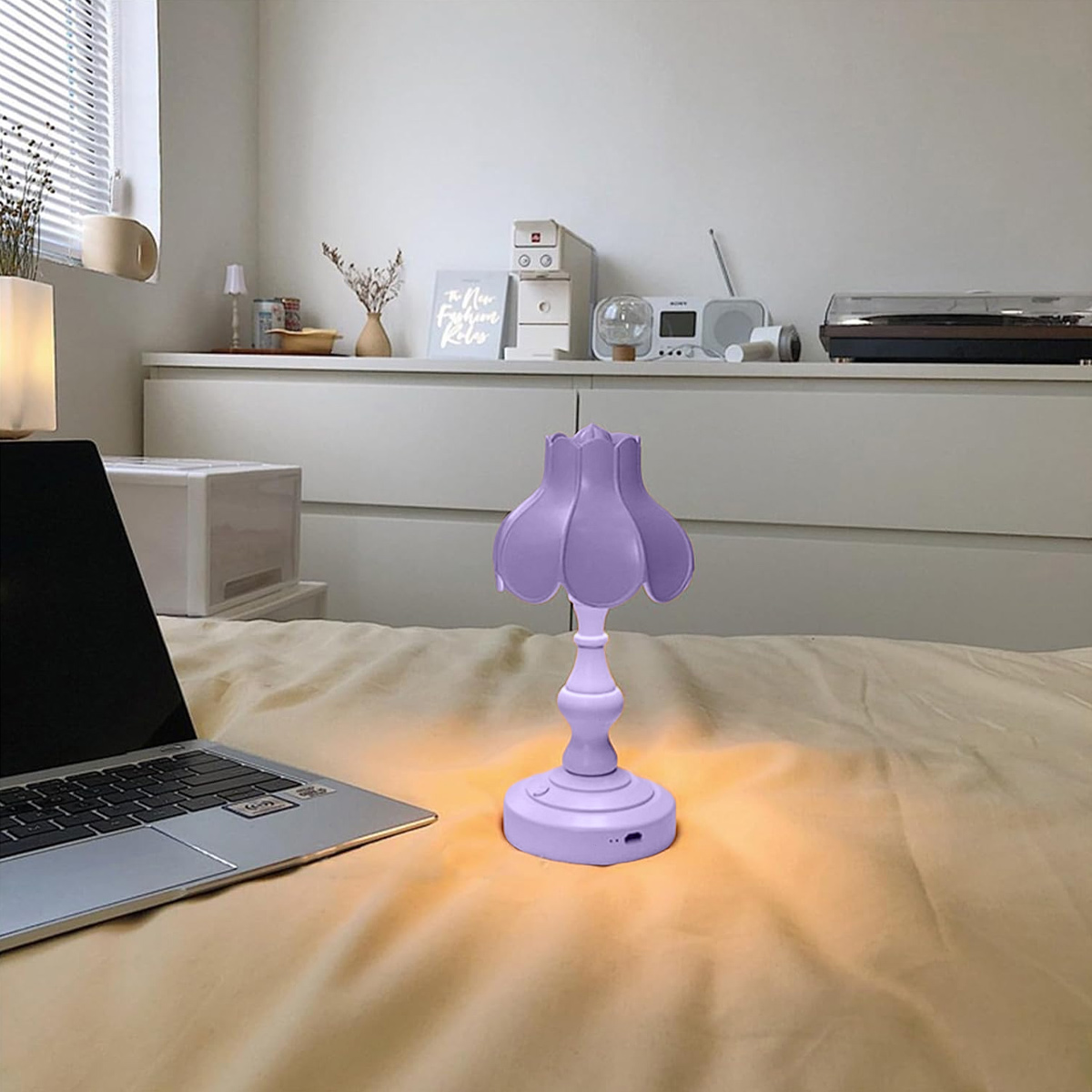 Eine lila Lampe auf einem Bett. Links steht ein Laptop. Im Hintergrund eine weiße Kommode mit Dekoration.