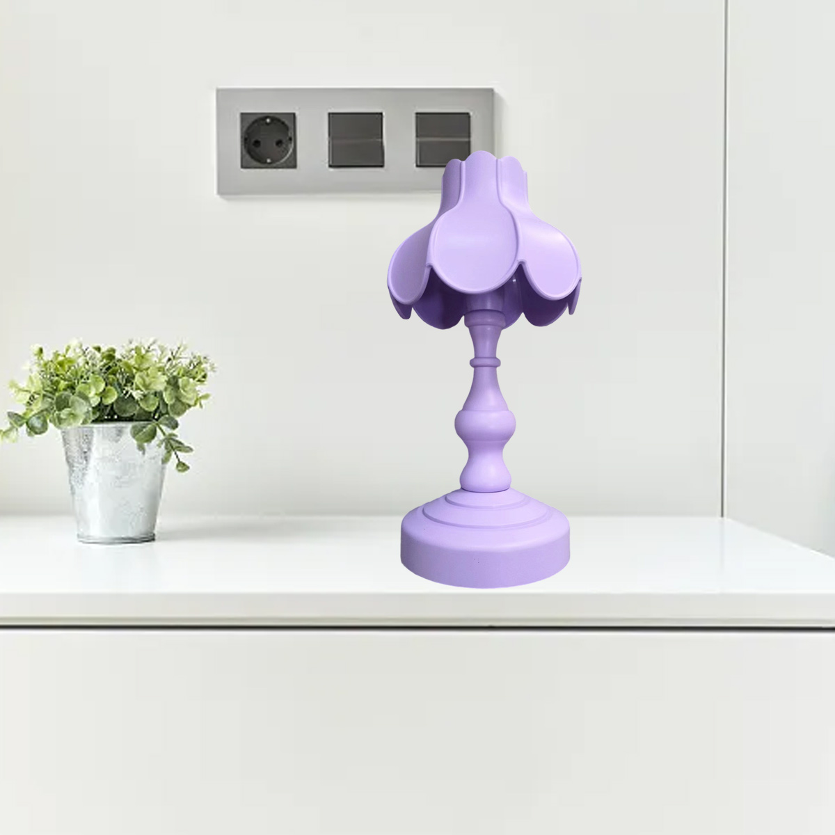 Eine lavendelfarbene Tischlampe steht auf einer weißen Oberfläche. Links ist eine kleine Pflanze in einem silbernen Topf. Steckdosen sind an der Wand.