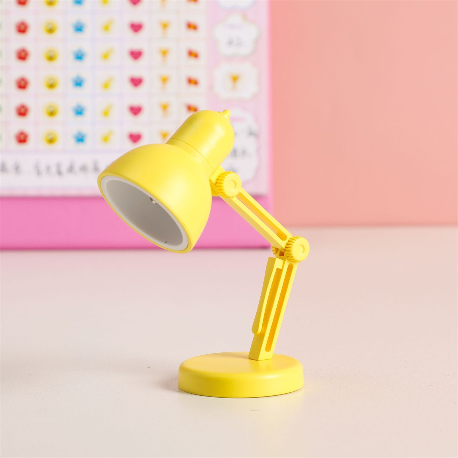 Eine gelbe Schreibtischlampe steht auf einer weißen Oberfläche. Ein rosa Hintergrund hat Aufkleber mit Herzen und anderen Formen.