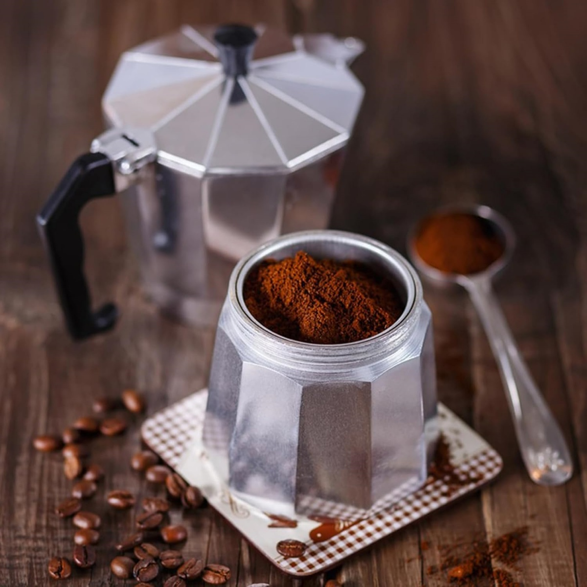 Eine Kaffeemaschine mit Kaffeepulver in der Mitte. Ein Löffel und einige Kaffeebohnen liegen ebenfalls auf dem Holztisch.