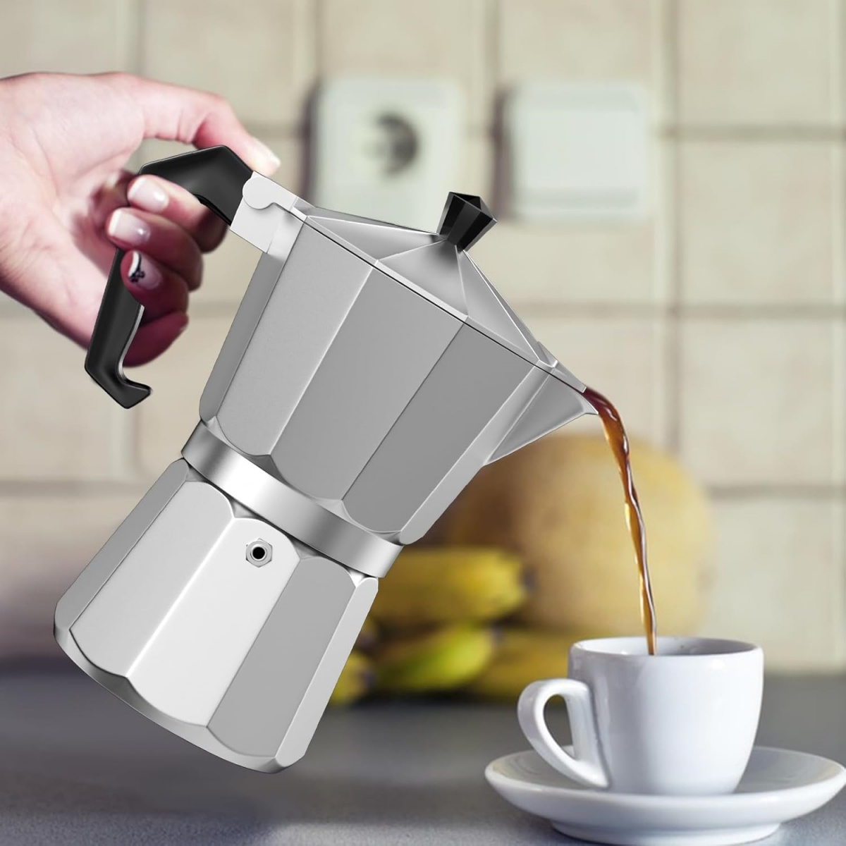 Eine Hand gießt Kaffee aus einer silbernen Espressomaschine in eine weiße Tasse auf einer Untertasse. Bananen und eine Melone sind im Hintergrund.