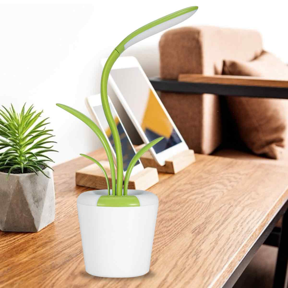 Eine Schreibtischlampe in Form einer Pflanze, weißer Sockel, grüner Stiel und Blätter. Zwei Telefone stehen im Hintergrund auf Holzständern. Links eine kleine Pflanze.