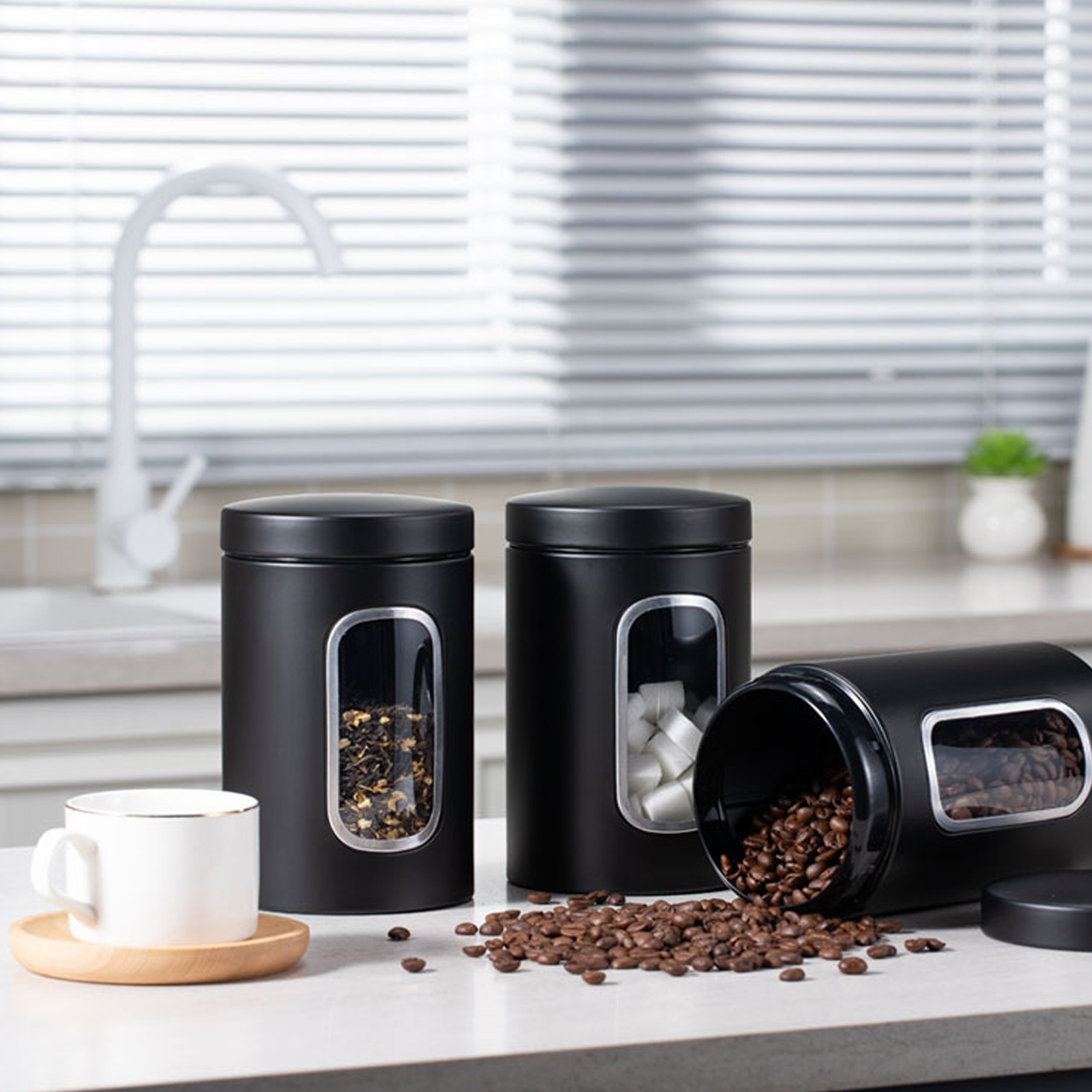 Schwarze Behälter mit Tee, Zucker und Kaffeebohnen. Eine Tasse und Untertasse stehen auf der Theke.