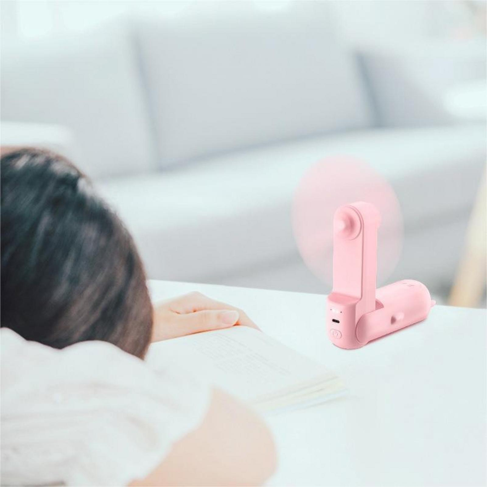 Eine Person beugt sich über ein Buch, auf dem Tisch steht ein rosa Ventilator. Im Hintergrund ist ein weißes Sofa zu sehen.