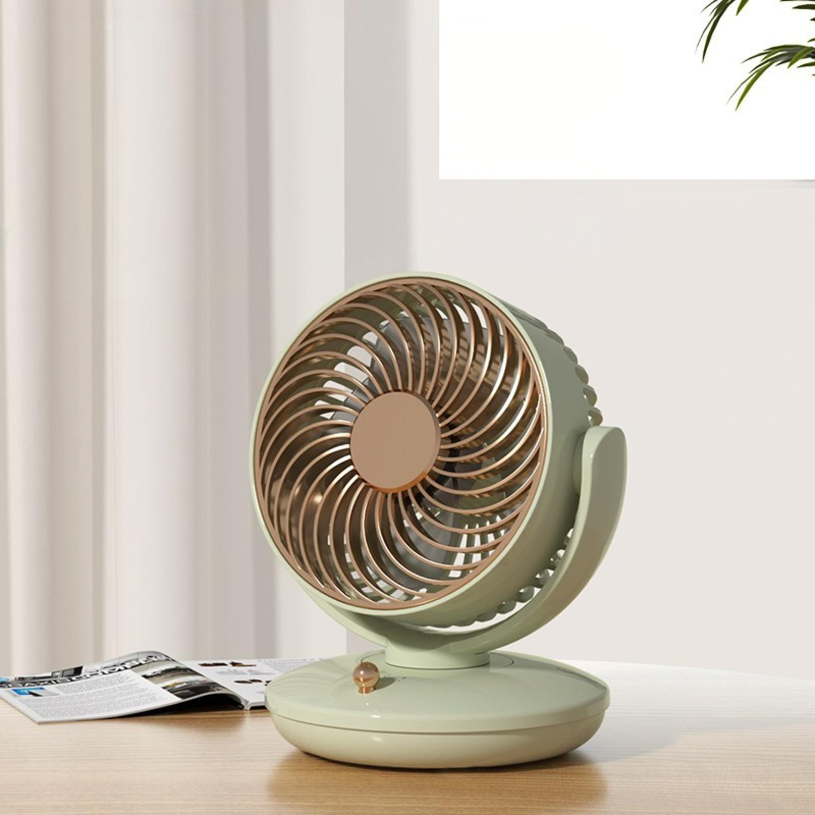 Ein mintgrüner und goldfarbener Ventilator steht auf einem Holztisch. Daneben liegt eine aufgeschlagene Zeitschrift. Vorhänge und ein Fenster sind zu sehen.