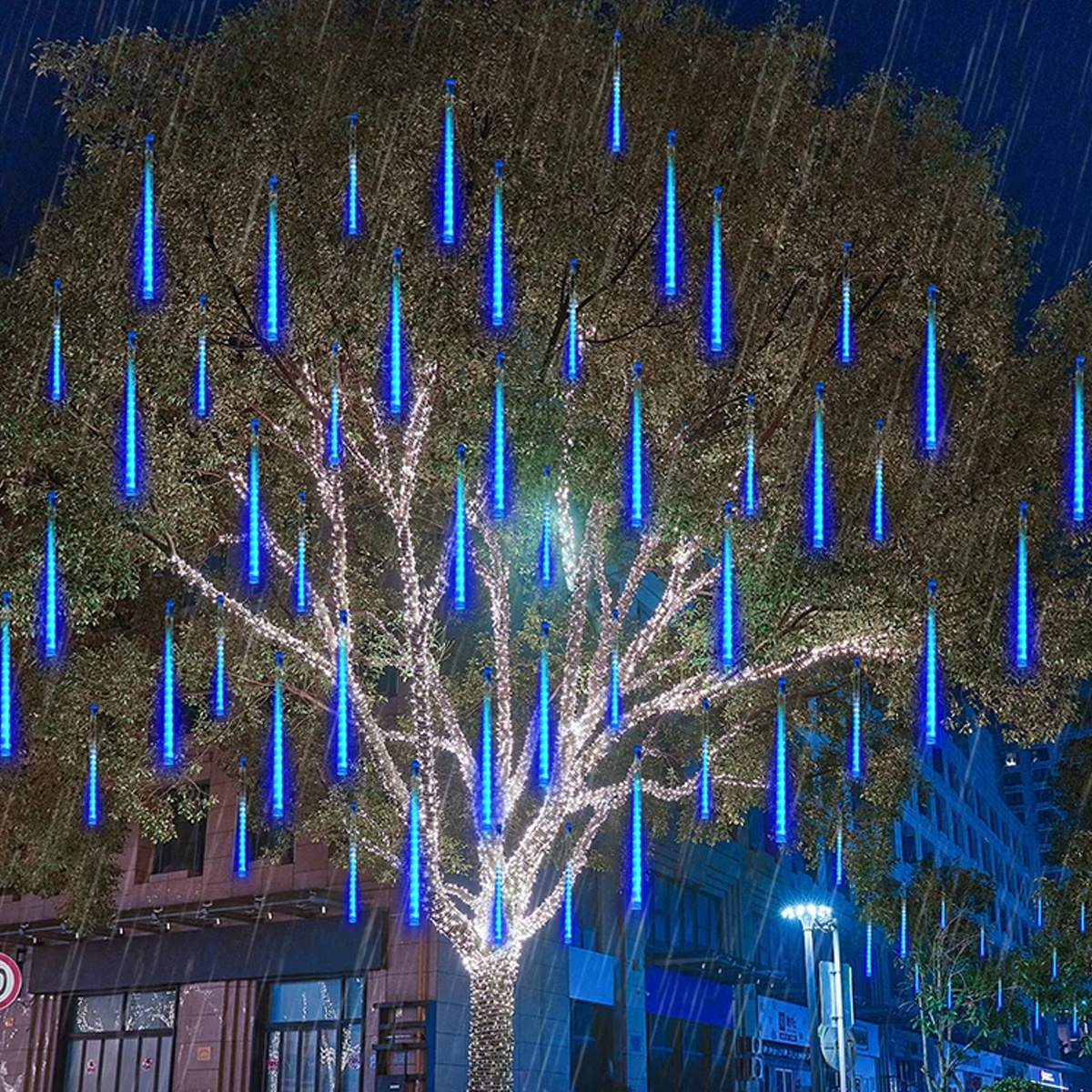Ein Baum mit blauen, eiszapfenförmigen Lichtern. Es regnet. Gebäude sind im Hintergrund. Es ist Nacht.