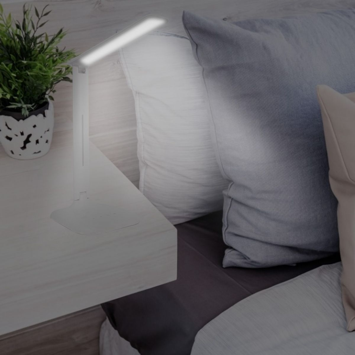 Eine weiße Schreibtischlampe leuchtet auf einem weißen Nachttisch. Kissen in Weiß- und Grautönen liegen auf einem Bett. Eine kleine Pflanze ist in einem Topf.