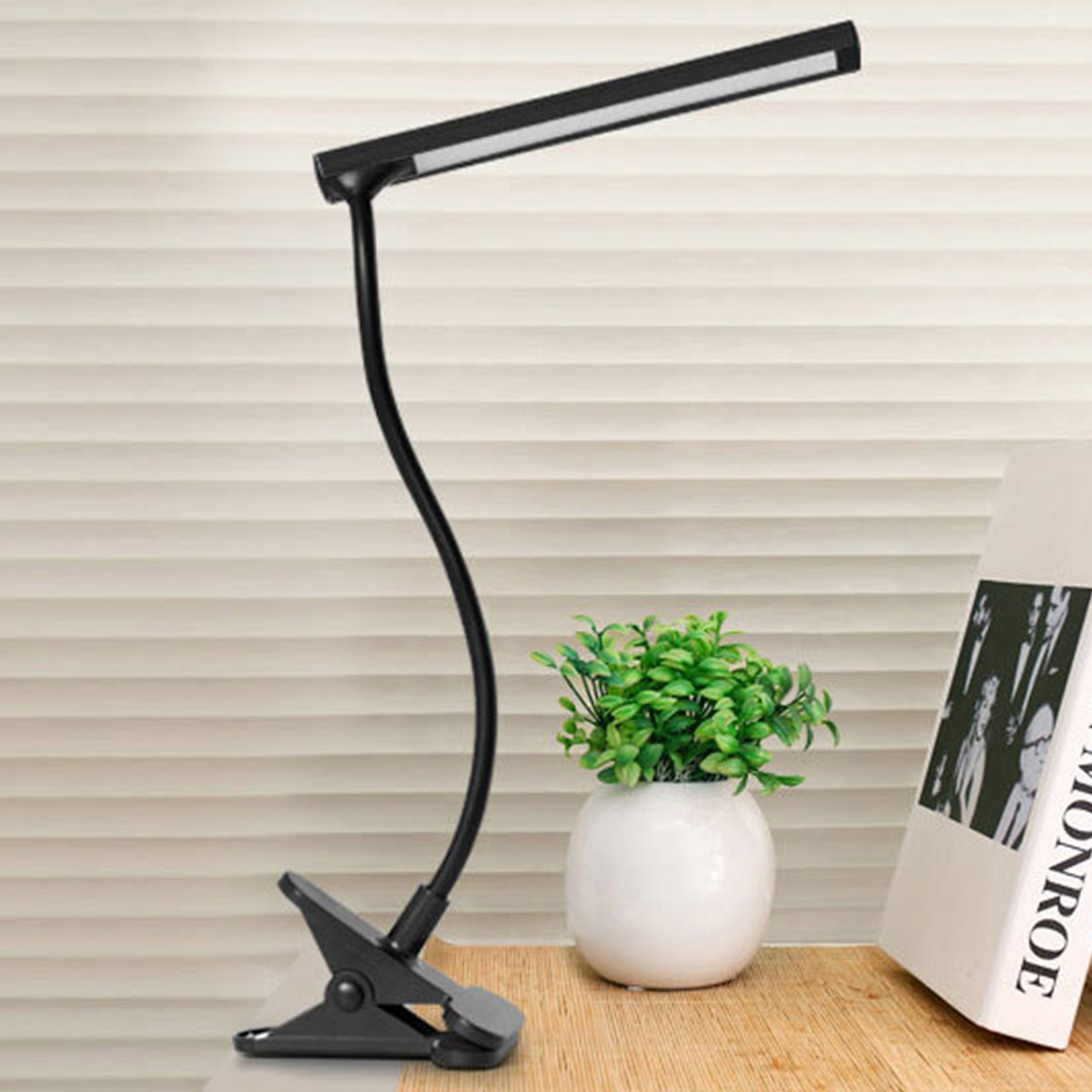 Eine schwarze Schreibtischlampe mit Clipfuß. Sie steht auf einer Holzoberfläche, neben einer weißen Vase mit grünen Pflanzen und einem Buch.