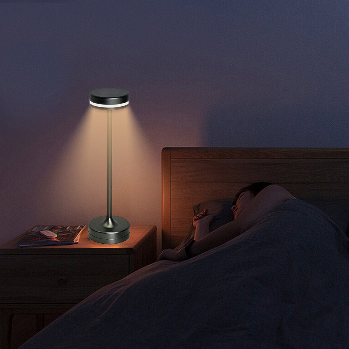 Eine Szene am Bett mit einer beleuchteten Lampe. Eine Person schläft im Bett. Ein Nachttisch mit Lampe und Zeitschriften.