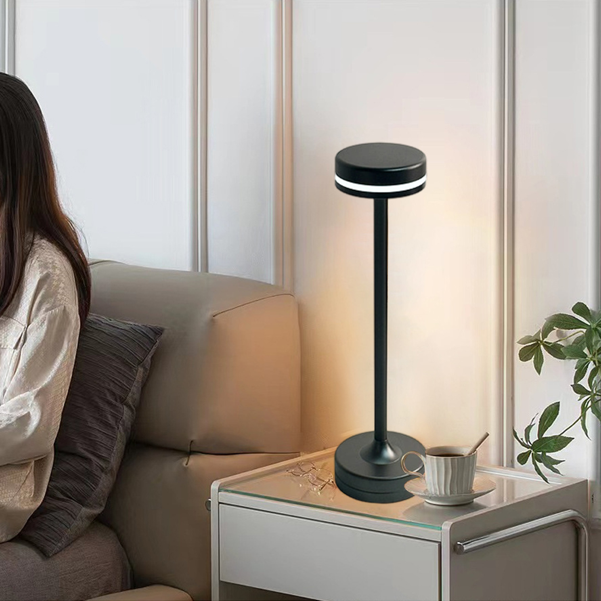 Eine moderne schwarze Lampe steht auf einem Nachttisch mit einer Kaffeetasse. Im Hintergrund sitzt eine Person auf einem Sofa.