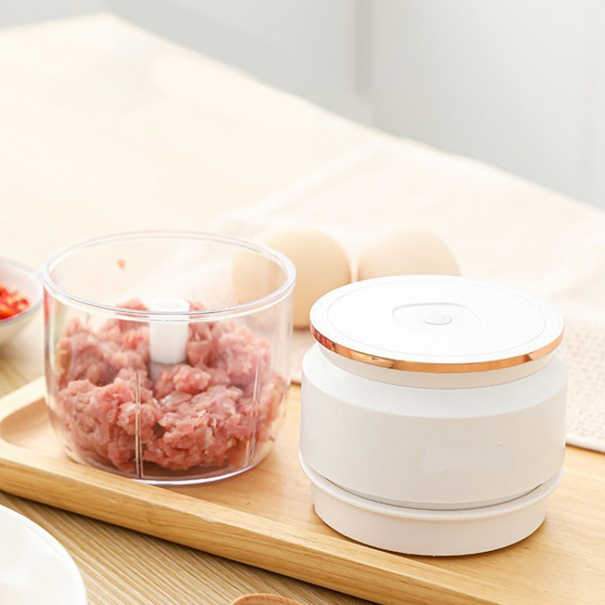 Ein Foodprozessor mit Hackfleisch, neben einem weißen Gerät und Eiern auf einem Holztablett auf einem Tisch.