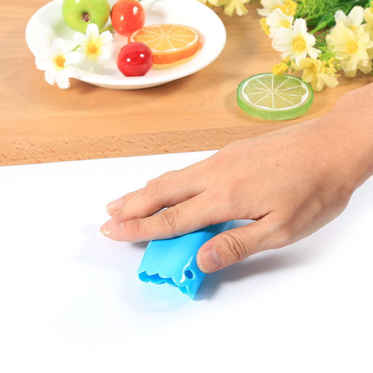 Eine Hand hält ein blaues Werkzeug. Früchte auf einem Teller und Blumen im Hintergrund. Weiße Oberfläche.