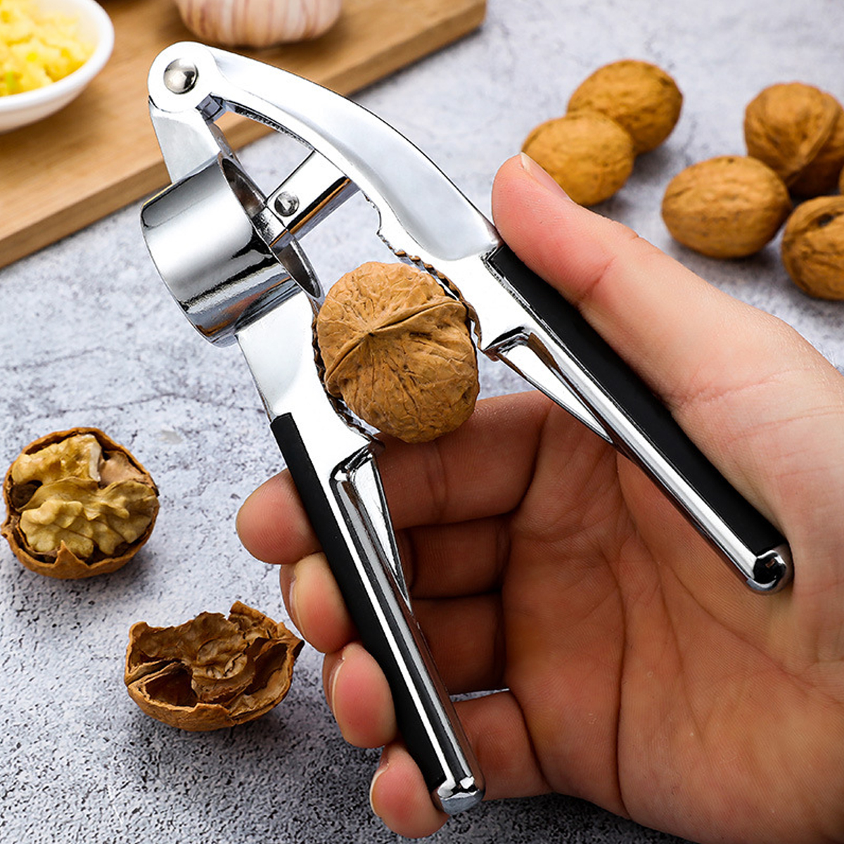 Hand hält eine Nussknacker mit einer Walnuss darin. Mehrere Walnüsse, Knoblauch und ein Holzbrett im Hintergrund.