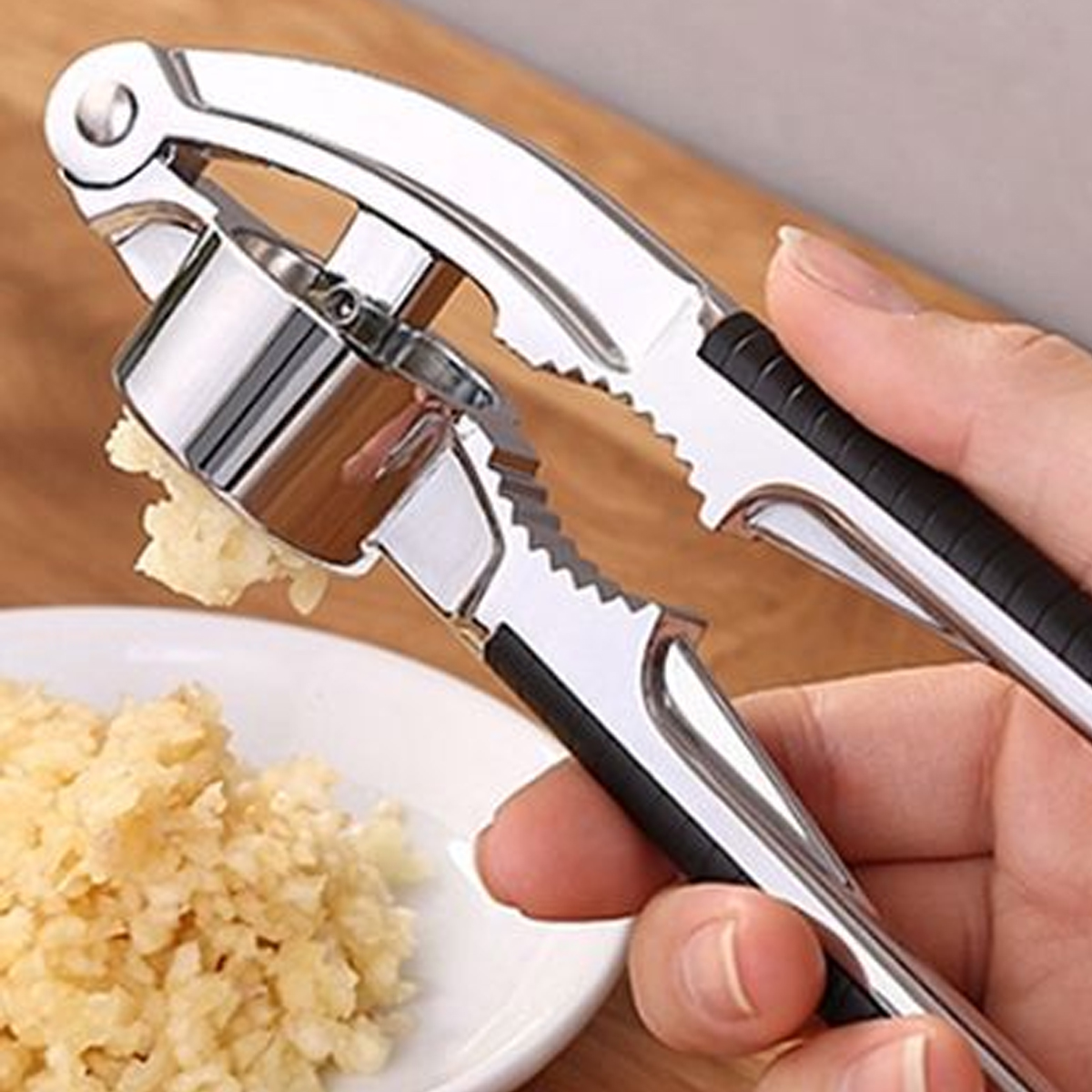 Eine Hand drückt eine Knoblauchpresse und extrudiert gehackten Knoblauch auf einen weißen Teller. Die Presse ist silber und schwarz.