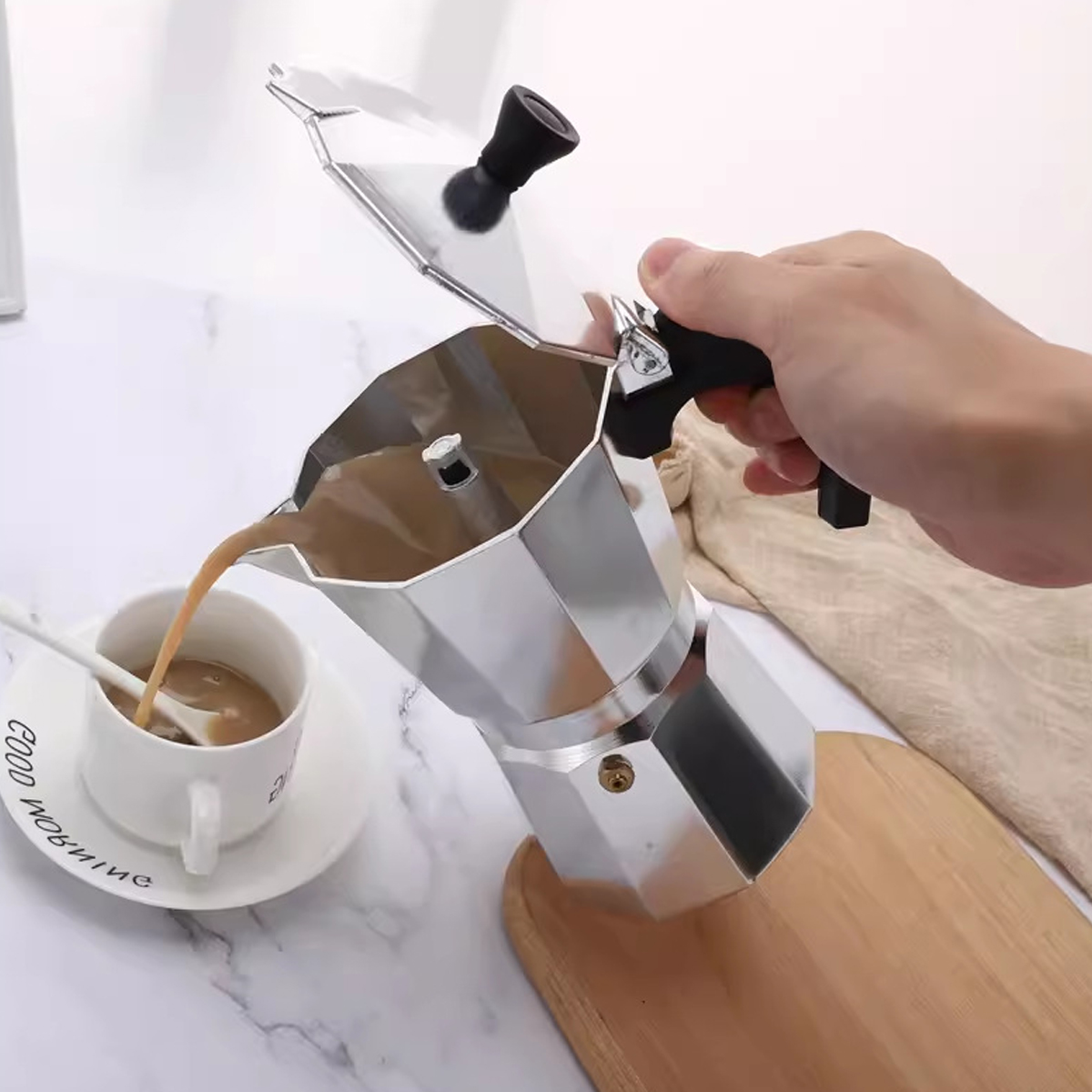 Eine Hand gießt Kaffee aus einer silbernen Moka-Kanne in eine weiße Tasse. Ein Löffel liegt in der Tasse auf einer Untertasse. Unten ist ein Holzbrett.