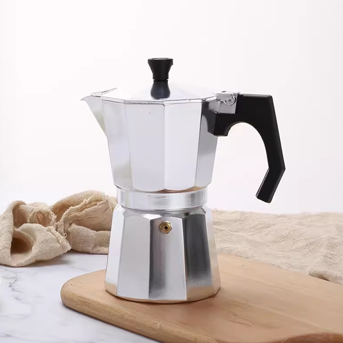 Eine silberne italienische Kaffeemaschine auf einem Holzbrett. Sie hat einen schwarzen Griff und Deckel. Beiges Tuch und weißer Hintergrund.