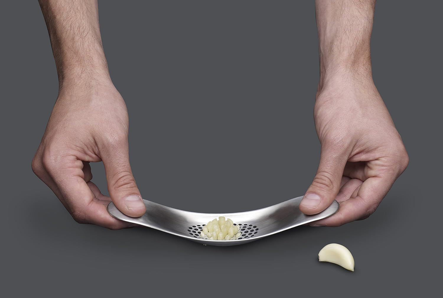 Hände halten eine Knoblauchpresse mit gepresstem Knoblauch. Eine Knoblauchzehe liegt daneben. Grauer Hintergrund.