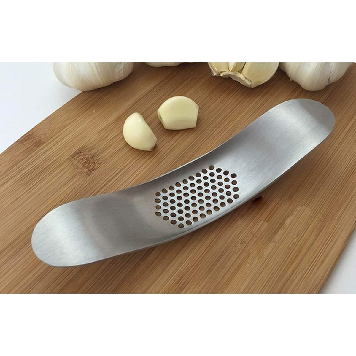 Eine Knoblauchpresse aus Metall mit Knoblauchzehen und -knollen auf einem Holzbrett. Die Presse hat viele kleine Löcher.