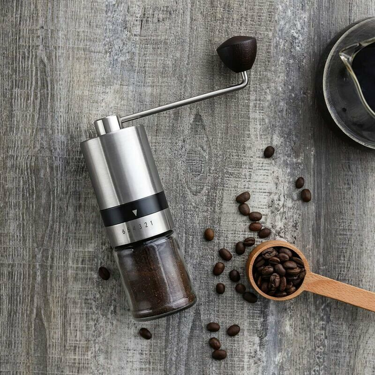 Eine Kaffeemühle mit Kaffeebohnen und einem Holzschöpfer auf einer Holzoberfläche. Eine Tasse mit Kaffee ist sichtbar.