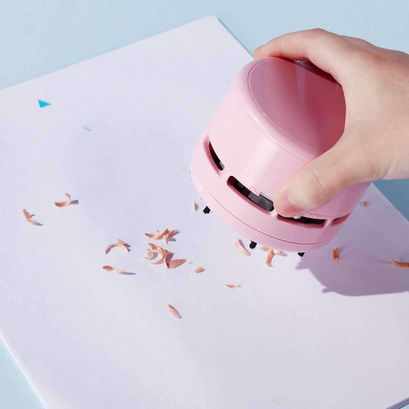 Eine Hand hält einen rosa Mini-Staubsauger, der Holzspäne von weißem Papier reinigt. Der Hintergrund ist hellblau.
