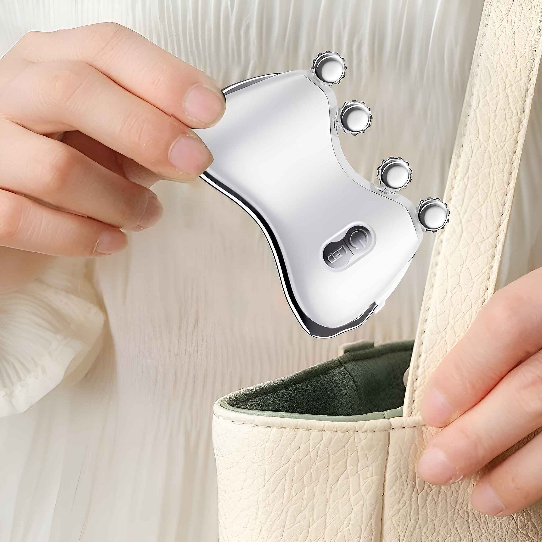 Eine Person hält ein silbernes Gesichtsmassagegerät über einer cremefarbenen Handtasche. Das Massagegerät hat runde Knöpfe.