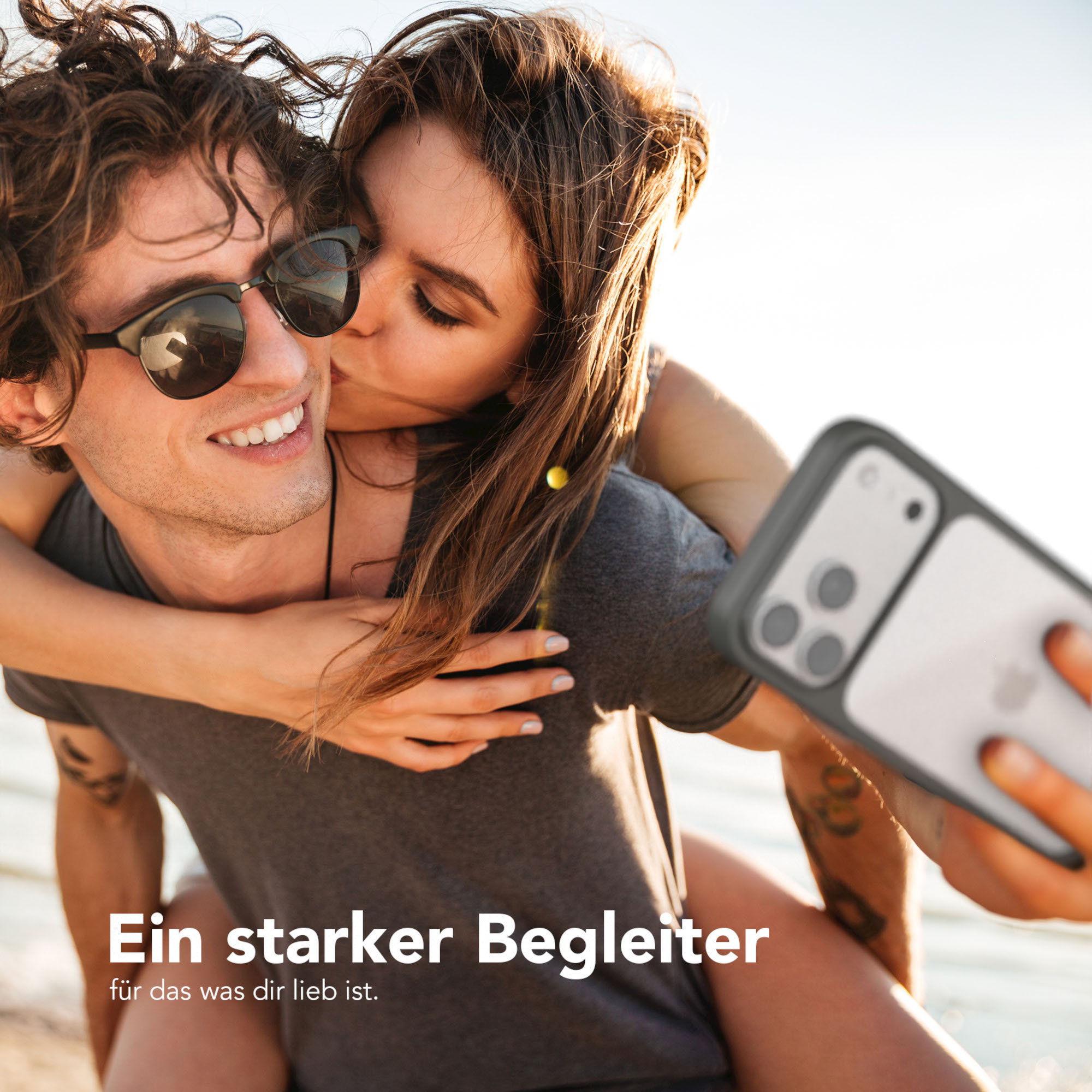 Ein Paar macht ein Selfie. Die Frau küsst die Wange des Mannes. Beide tragen Sonnenbrillen. Das Telefon hat eine Hülle.