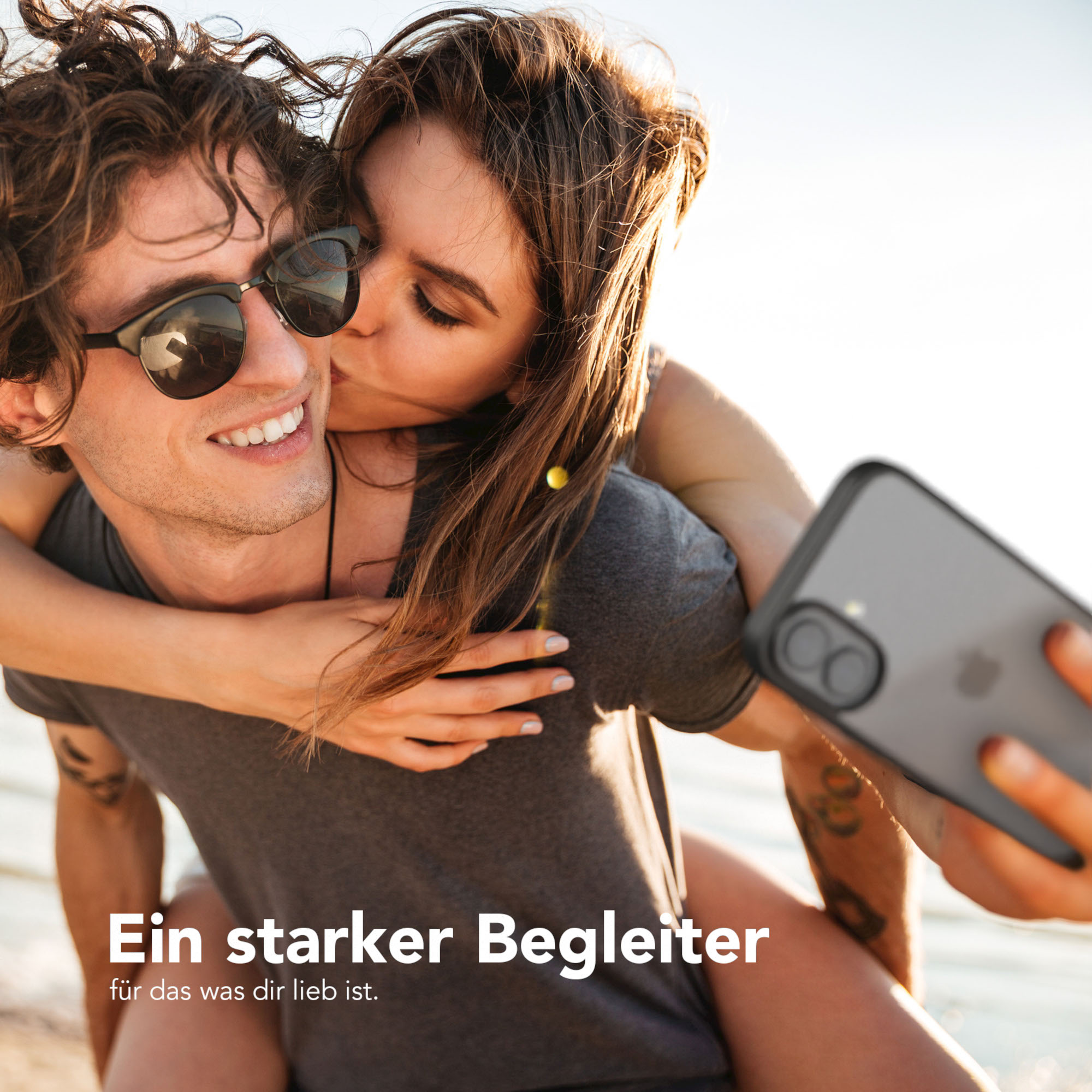 Ein Mann lächelt, während er von einer Frau von hinten geküsst wird und ein Selfie mit einem Telefon macht. Der Hintergrund ist das Meer.