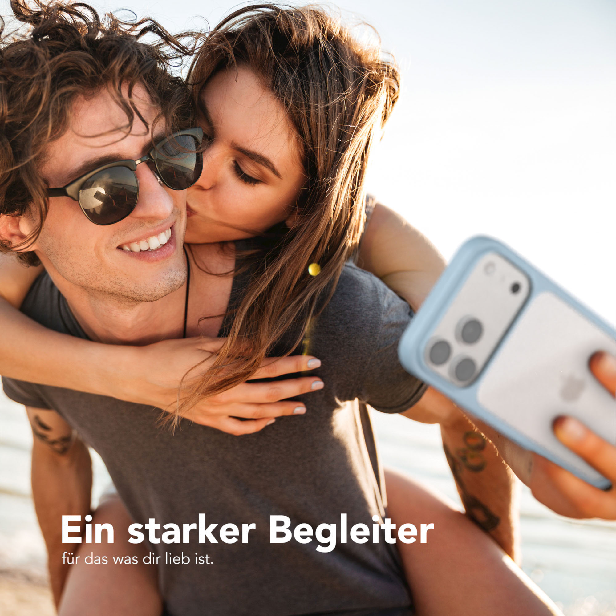 Eine Frau küsst einen Mann auf die Wange. Er trägt eine Sonnenbrille. Sie machen ein Selfie mit einem Telefon.