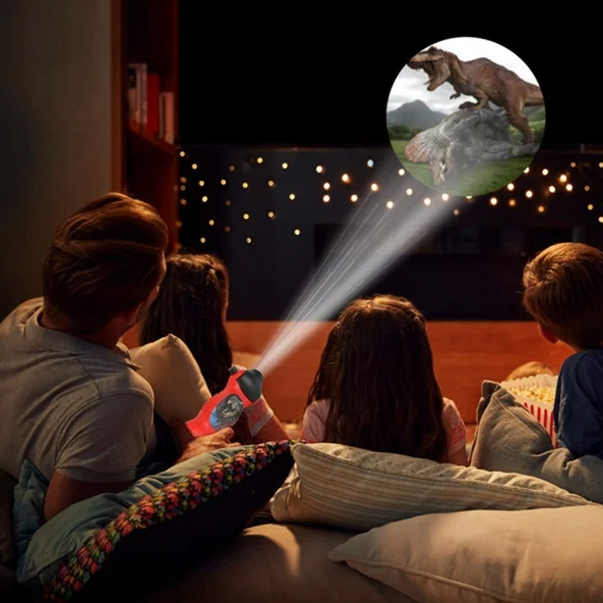 Eine Familie sieht eine Dinosaurierprojektion. Ein roter Projektor wirft Licht auf eine Leinwand mit einem T-Rex auf einem Felsen. Kinder schauen zu.