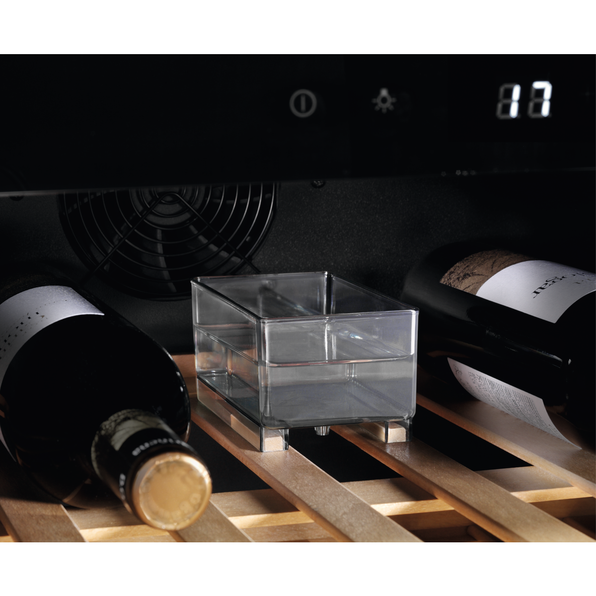 Dentro de una vinoteca: botellas, recipiente transparente, estantes de madera. Pantalla digital muestra 87. Ventilador negro.