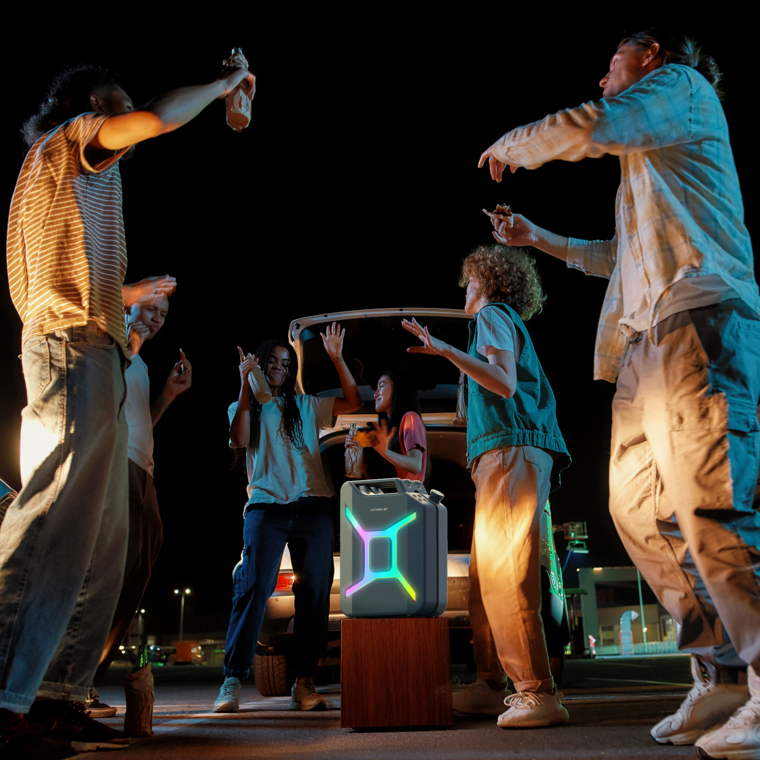 Grupa młodych ludzi tańczących wokół głośnika w nocy. Niektórzy trzymają drinki. Samochód w tle z otwartym bagażnikiem. Neonowe światła.