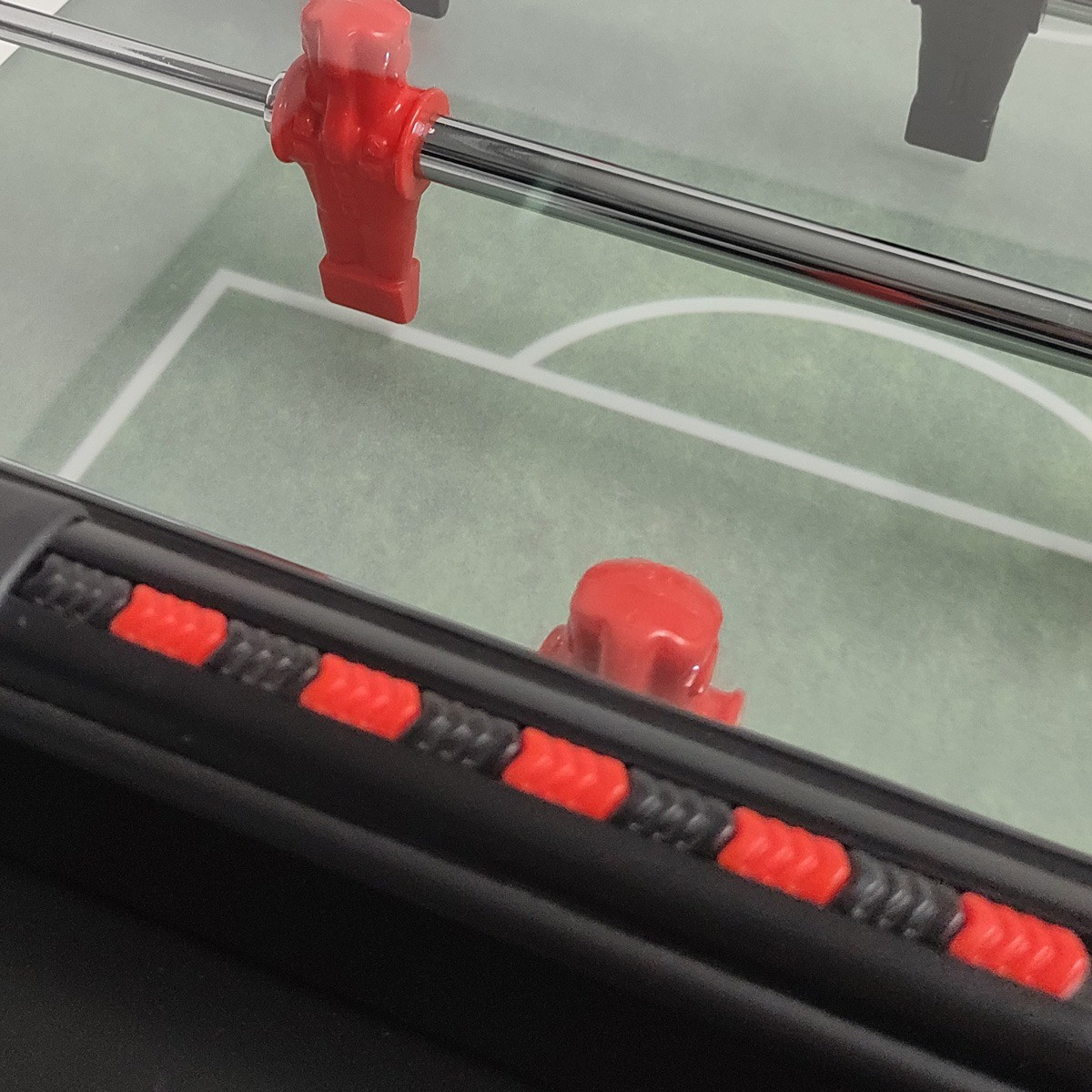 Close-up van een tafelvoetbaltafel. Rode spelersfiguren op staven, zwart en rode accenten. Groen speelveld met witte lijnen.