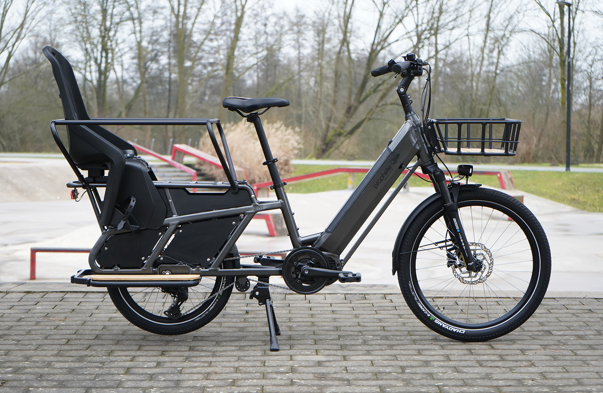 Ein graues Cargobike mit schwarzem Kindersitz steht auf einer Backsteinoberfläche. Bäume und ein Skatepark sind im Hintergrund.