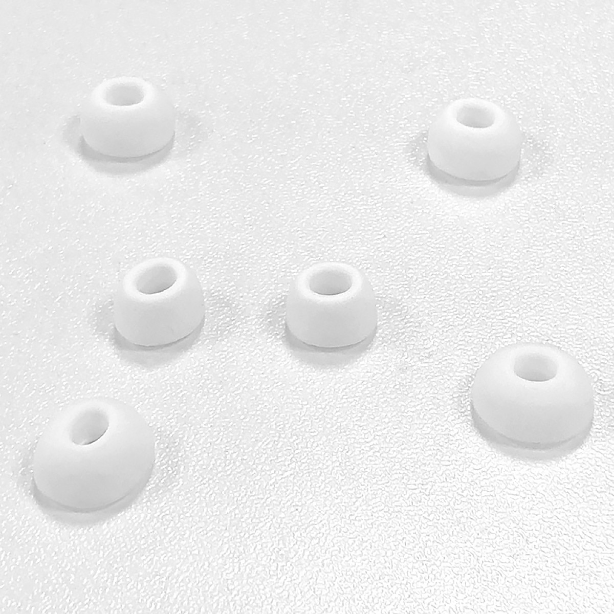 Sześć białych końcówek do słuchawek na teksturowanej białej powierzchni. Każda końcówka to zaokrąglony walec z centralnym otworem.