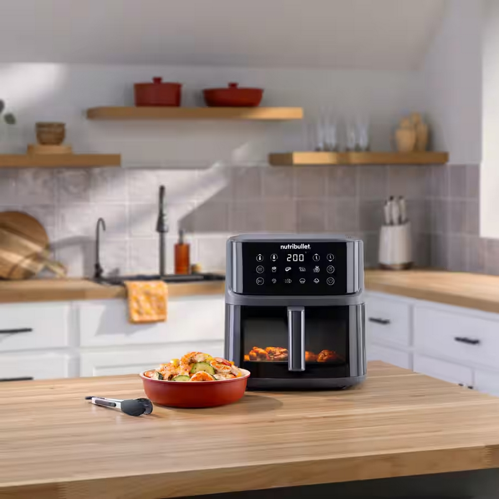 Scena kuchenna z frytkownicą Nutribullet, miską jedzenia i przyborami kuchennymi na drewnianym blacie.
