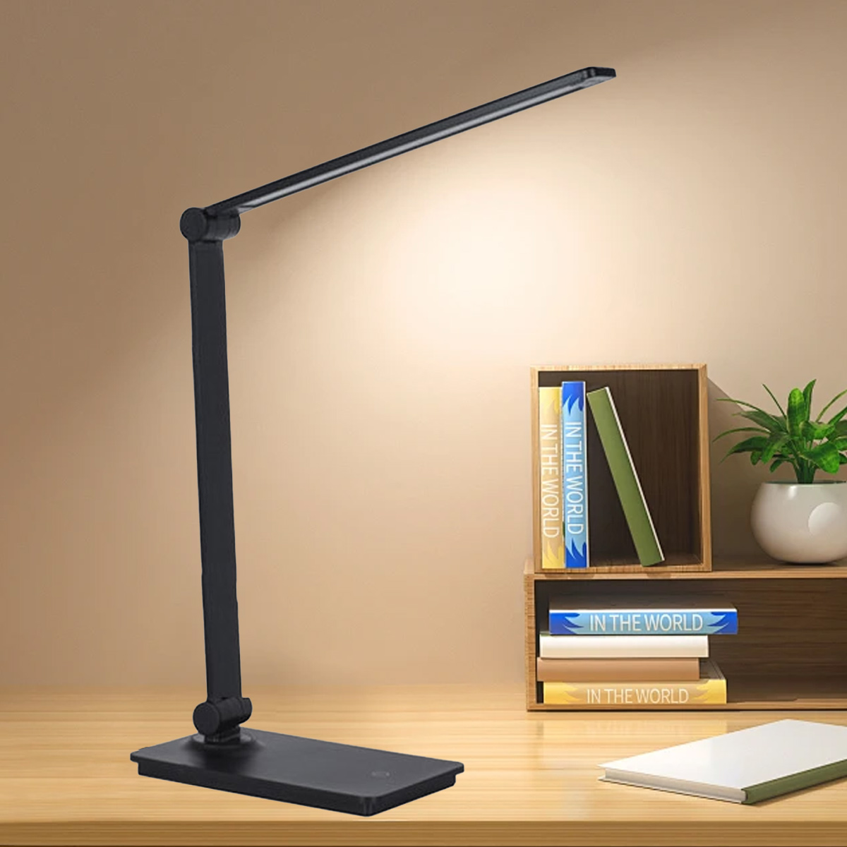 Eine schwarze Schreibtischlampe steht auf einem Holztisch. Bücher und eine Pflanze befinden sich im Hintergrund. Die Lampe leuchtet.