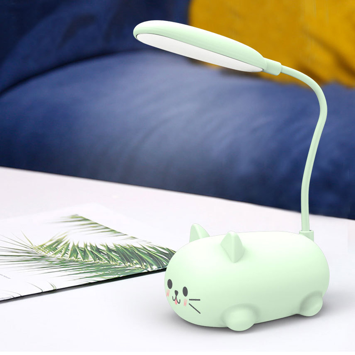 Eine mintgrüne, katzenförmige Schreibtischlampe mit flexiblem Hals und weißem Licht. Sie steht auf einem weißen Tisch mit Pflanzenillustration.