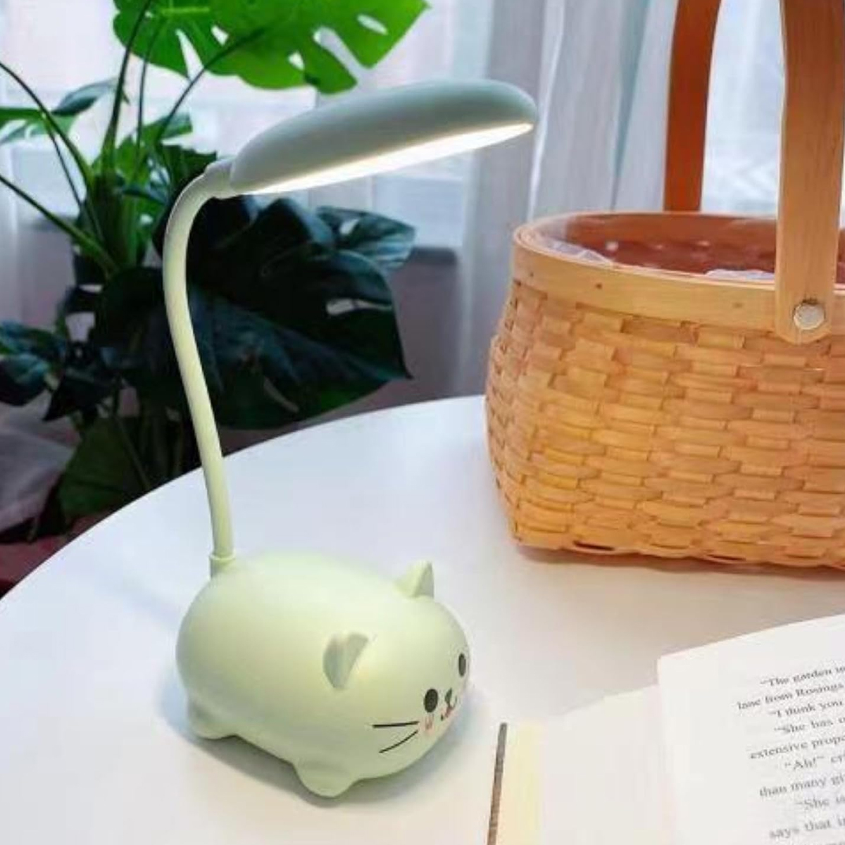 Eine katzenförmige Schreibtischlampe steht auf einem weißen Tisch, neben einem Korb und einem Buch. Grüner Pflanze im Hintergrund.
