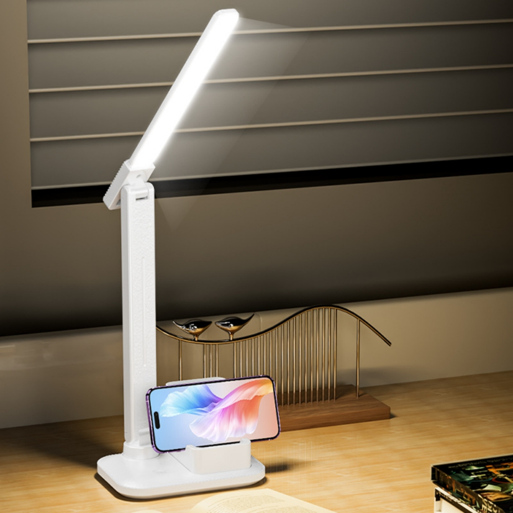 Weiße Schreibtischlampe mit Telefonaufladung auf der Basis. Das Telefon zeigt ein farbenfrohes Bild. Hintergrund zeigt dekorative Gegenstände.