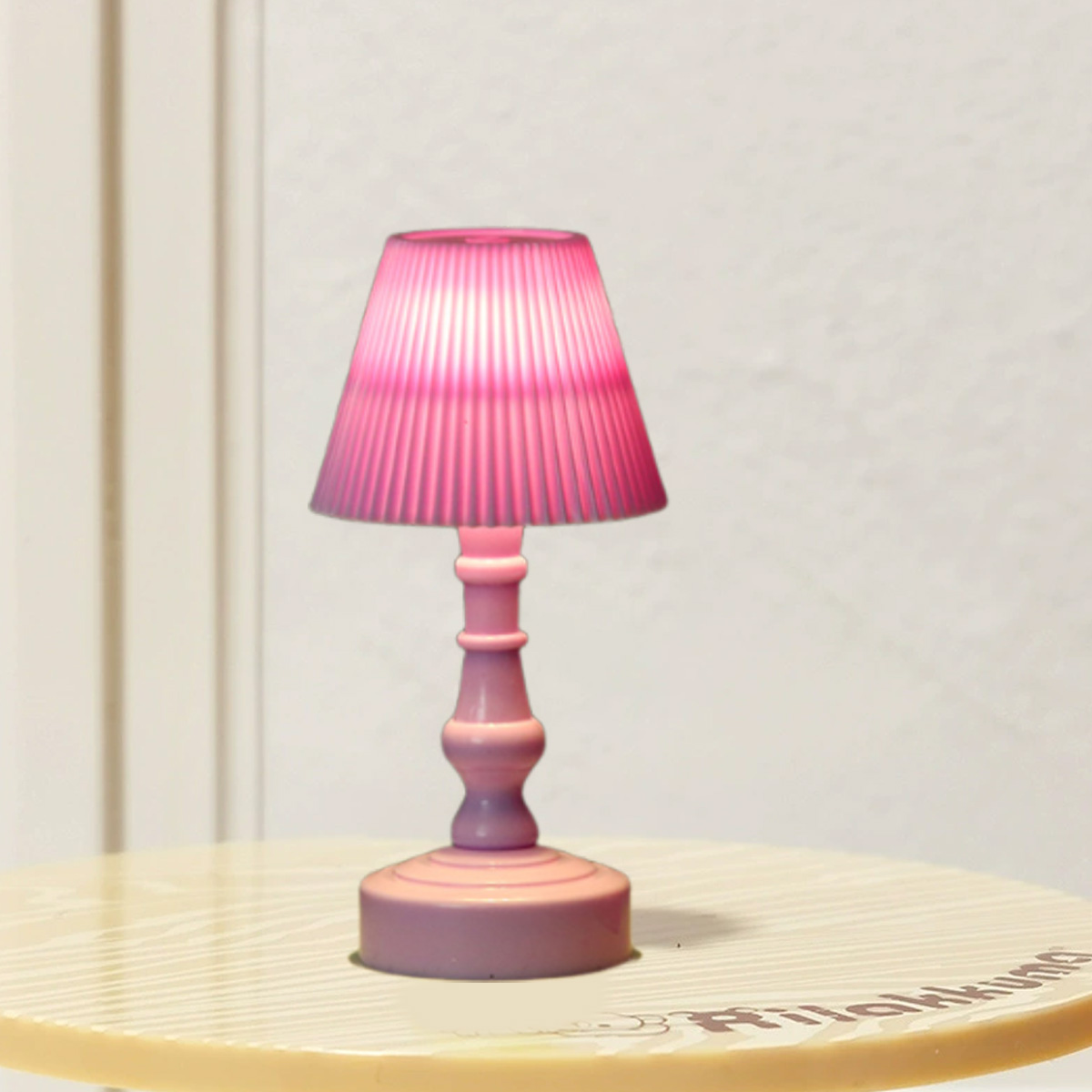 Eine rosa Tischlampe mit plissiertem Lampenschirm steht auf einem Tisch. Der Hintergrund ist eine weiße Wand und ein hellfarbiger Tisch.