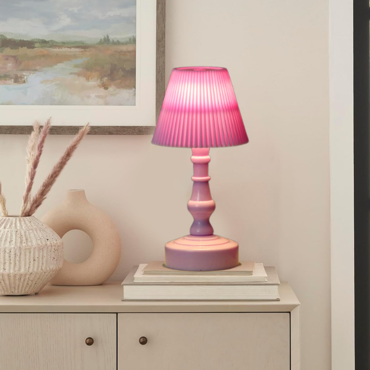 Eine rosa Lampe mit plissiertem Schirm steht auf Büchern. Daneben Vasen und Kunstwerk auf einem hellen Schrank.
