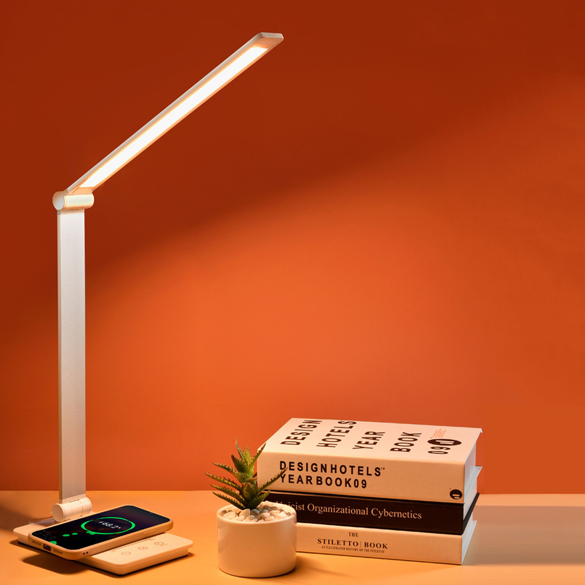 Eine weiße LED-Schreibtischlampe beleuchtet einen Schreibtisch mit Büchern, einer kleinen Pflanze und einem ladenden Telefon. Orangen Wand im Hintergrund.