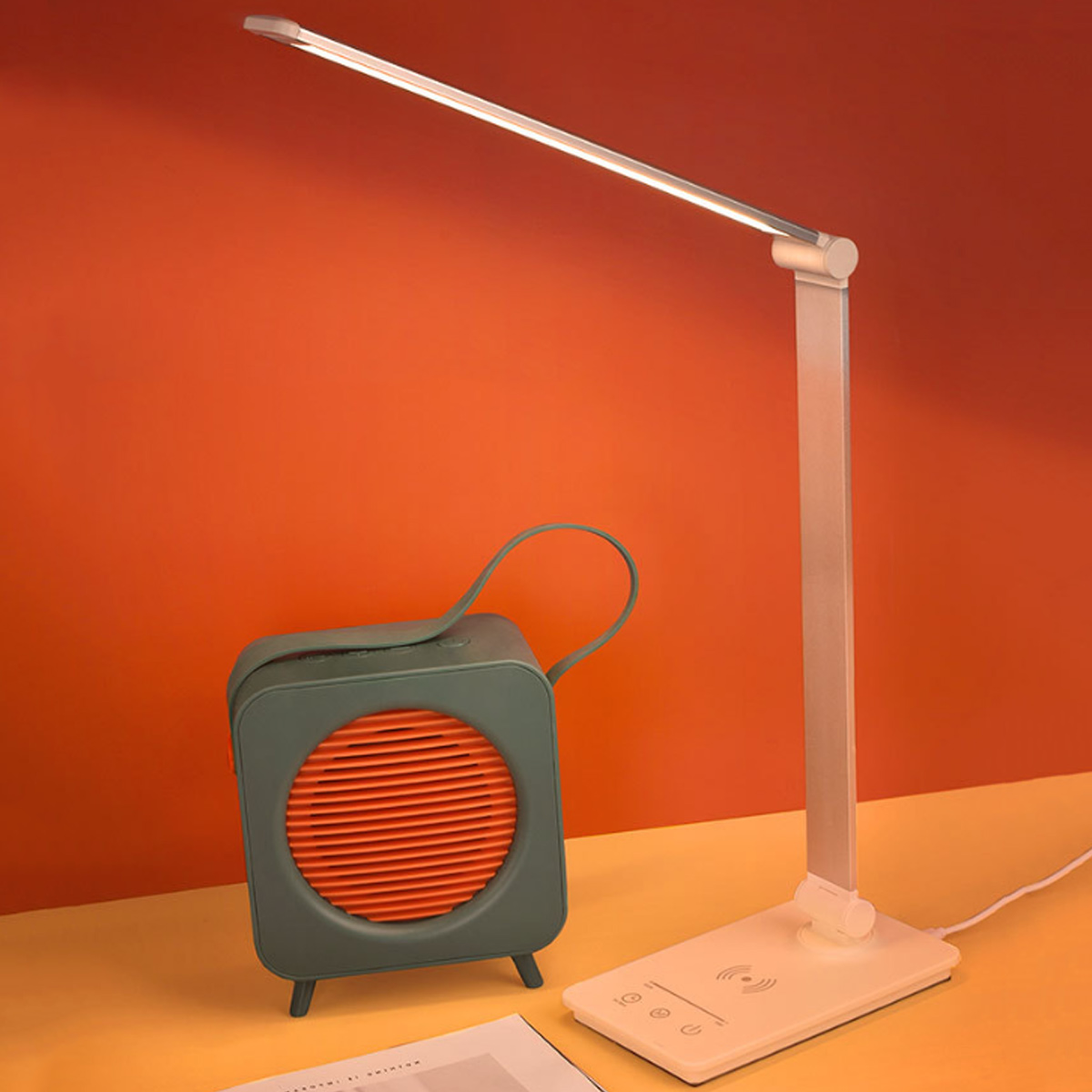Eine Schreibtischlampe und ein Retro-Lautsprecher stehen auf einem Schreibtisch vor einer orangefarbenen Wand. Die Lampe ist weiß und der Lautsprecher türkis und orange.
